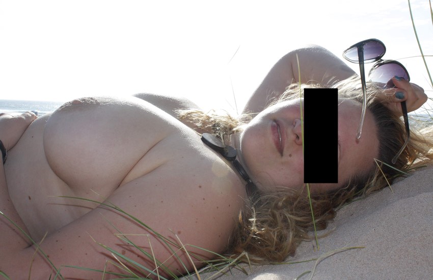 Une jolie femme forte allongée sur le sable d'une plage. Elle enlève ses lunettes de soleil, sa main sur le ventre elle une belle poitrine nue